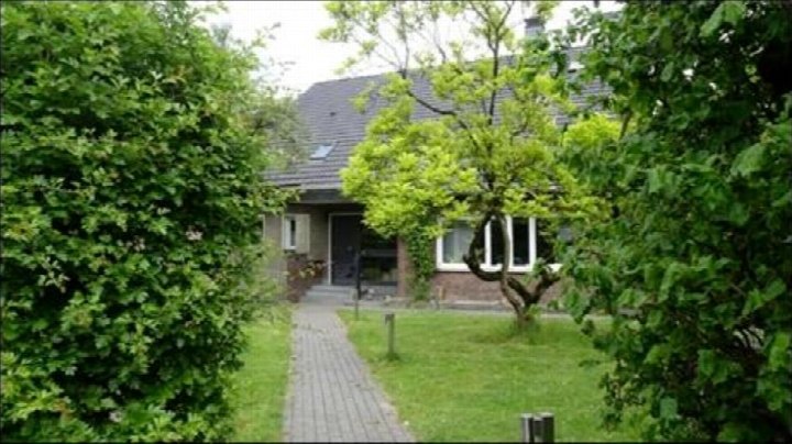 Vorderansicht eines Zweifamilienhauses mit Eingang, gepflastertem Weg und Vorgarten, umgeben von Bäumen und Sträuchern