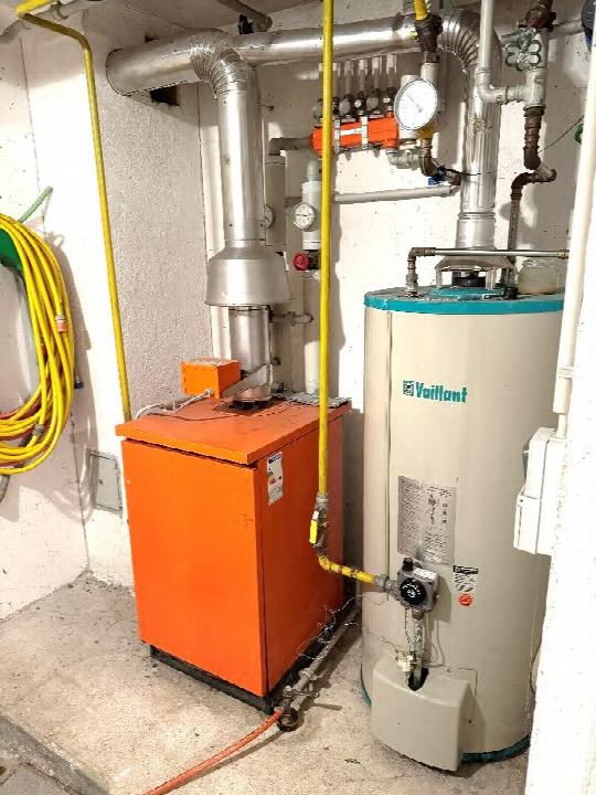 Technischer Raum mit orangefarbenem Heizkessel, Vaillant-Warmwasserboiler und Rohrleitungen in einem Kellerbereich