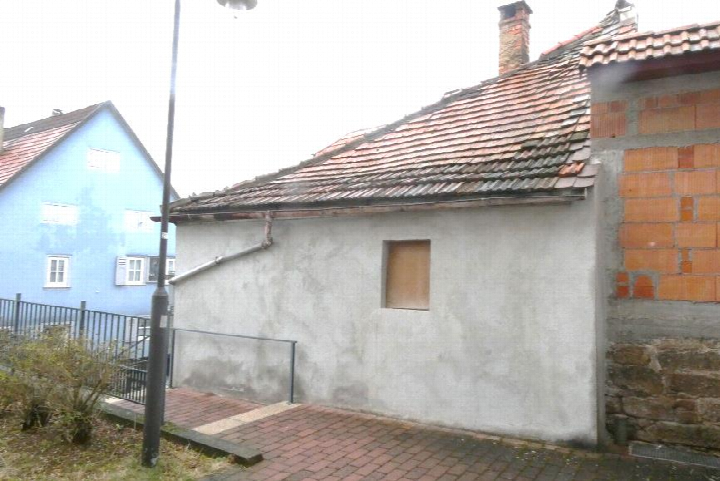Seitenansicht eines Gebäudes mit grauer Putzfassade, Ziegeldach und Schornstein; im Hintergrund ein blauer Anbau und ein Metallgeländer