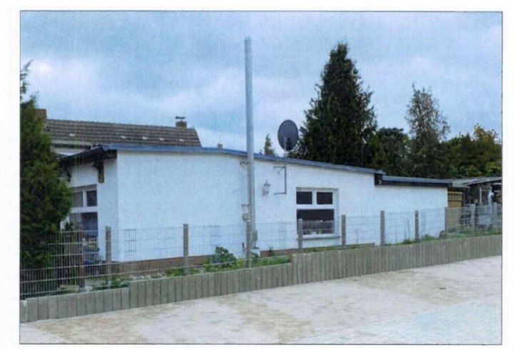 Seitenansicht eines Einfamilienhauses mit weißer Fassade, Fenstern, Satellitenschüssel und Metallzaun, umgeben von Bäumen und Grünanlagen