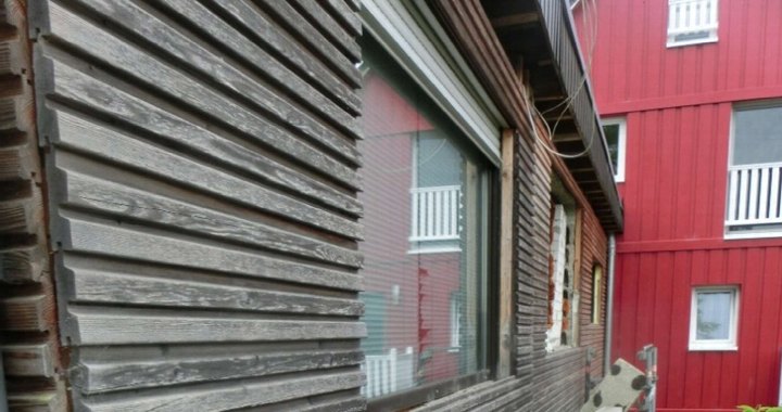 Seitenansicht eines Einfamilienhauses mit verwittertem Holzschindelverkleidung und großem Fenster, daneben ein rotes Gebäude