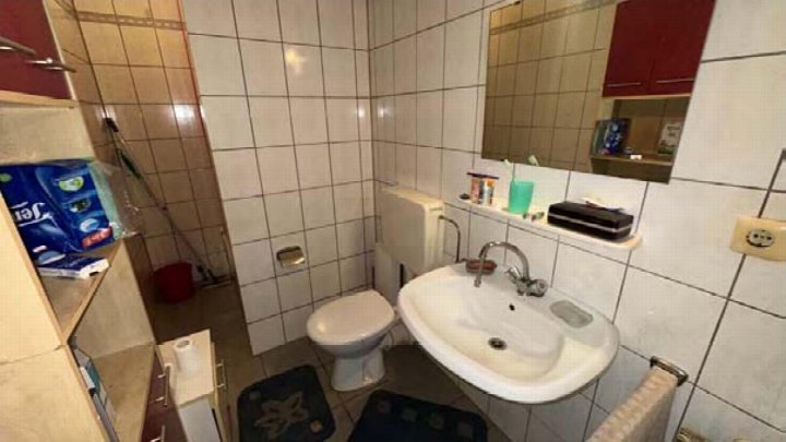 Badezimmer mit weiß gefliesten Wänden, Waschbecken, Toilette, Spiegel und Regal. Im Hintergrund Dusche und Reinigungsutensilien