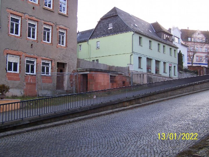 Seitenansicht eines Gebäudes mit grauer Fassade und roten Ziegelrahmen, daneben ein grünes Haus, gepflasterte Straße und Metallgeländer in Glauchau