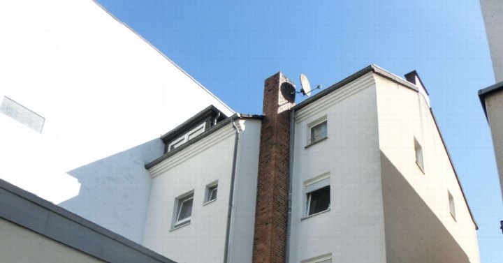 Seitenansicht eines Mehrfamilienhauses mit weißer Fassade, Fenstern, Ziegelkamin und Satellitenschüssel unter blauem Himmel