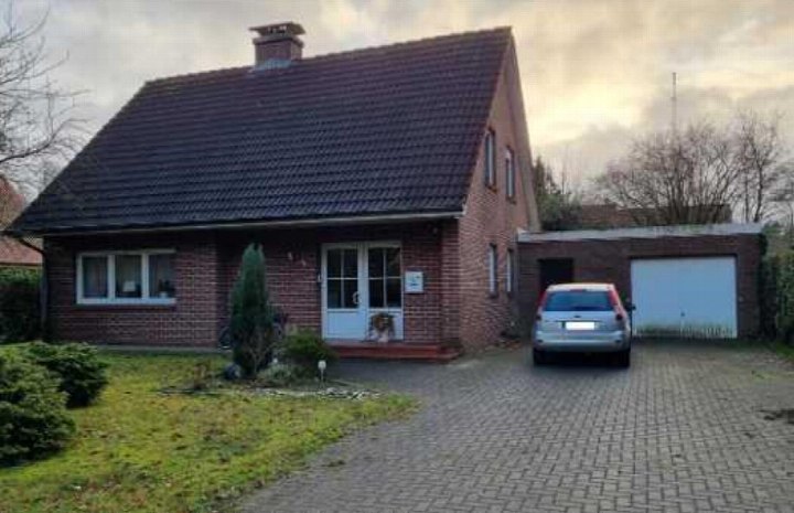 Einfamilienhaus aus Backstein mit Satteldach, Eingangstür, Fenstern und angrenzender Garage. Vor dem Haus befindet sich ein gepflasterter Vorplatz mit einem geparkten Auto und