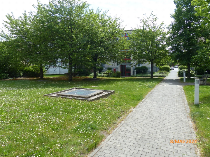 Grüner Gartenbereich mit Bäumen, Rasen, einem gepflasterten Weg und einem quadratischen Element im Vordergrund. Im Hintergrund ist ein Teil eines Gebäudes sichtbar