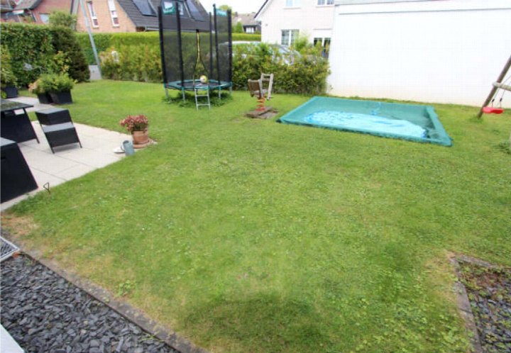Garten mit Rasenfläche, Trampolin, Sandkasten, Gartenmöbeln und Pflanzen; im Hintergrund Hecke und Nachbarhäuser