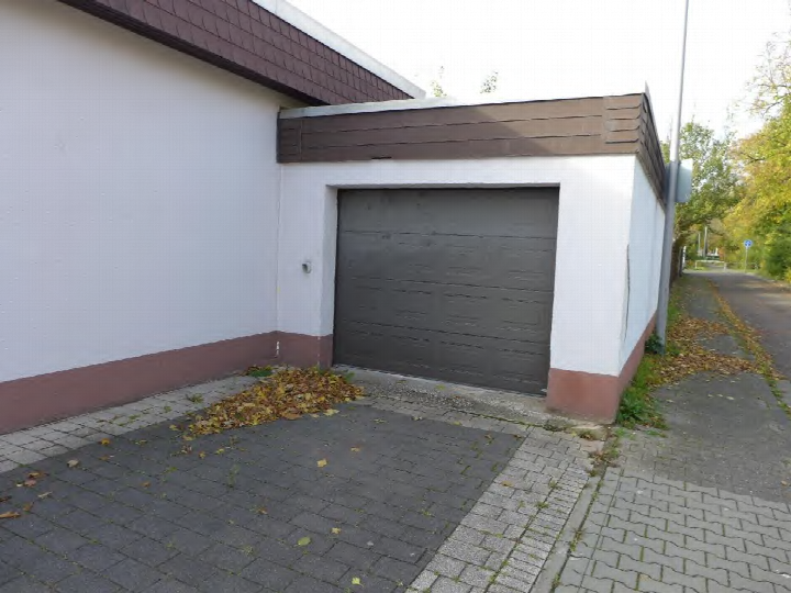 Garage mit dunkler Rolltür an einem weißen Gebäude, gepflasterter Zugang, herbstliche Laubablagerung