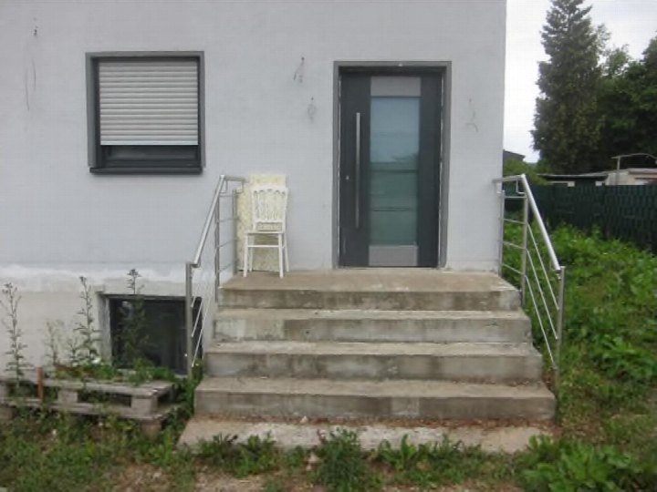Eingangsbereich eines Gebäudes mit Betontreppen, Metallgeländern, einer Glastür und einem Fenster mit Rollladen. Umgebung mit Grünflächen