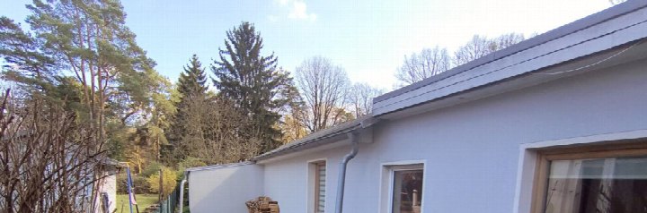 Seitenansicht eines Einfamilienhauses mit grauer Fassade, Dachrinne und Fenstern, im Hintergrund Bäume und Garten
