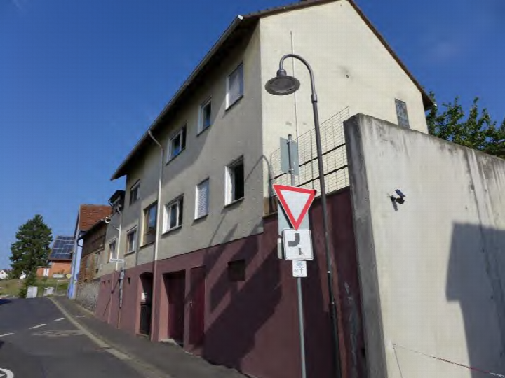 Seitenansicht eines zweistöckigen Wohnhauses mit grauer Fassade und rotem Sockelbereich, Straßenlaterne und Verkehrszeichen am Rand einer Straße in Bad Nauheim