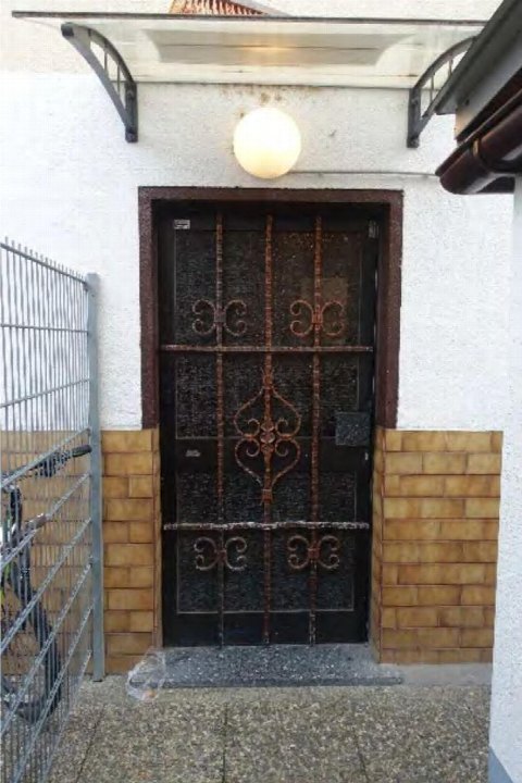 Eingangstür mit Metallgitter, darüber runder Außenleuchte, an weißer Fassade mit gelben Ziegelverkleidungen am Sockelbereich