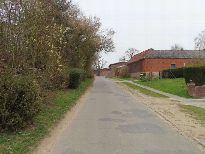 Seitenansicht eines roten Backsteinhauses an einer asphaltierten Straße mit Grünanlagen und Bäumen im Vordergrund