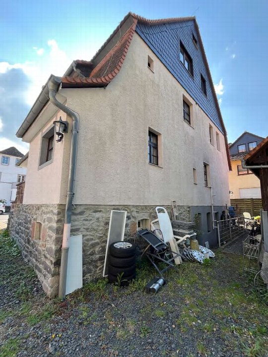 Seitenansicht eines Zweifamilienhauses mit Steinsockel, verputzter Fassade, Dach mit roten Ziegeln und blauen Schieferplatten sowie unordentlichem Gartenbereich
