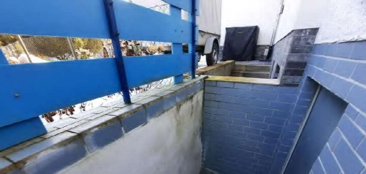 Blauer Metallzaun und Ziegelmauer eines Garagenbereichs mit Betonrampe und teilweise sichtbarem Lkw im Hintergrund