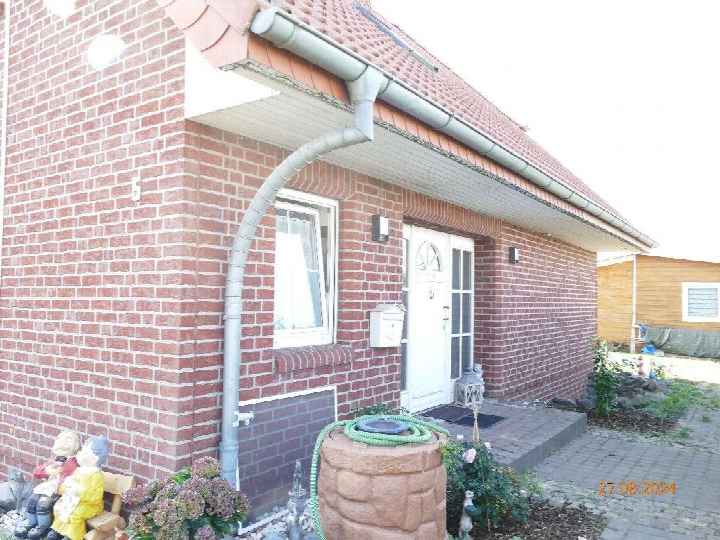 Eingangsbereich eines Backsteinhauses mit weißer Tür, Fenster und Regenrohr; im Vordergrund Dekoration und Gartenelemente