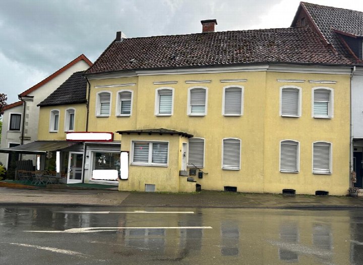 Gelbes Wohn- und Geschäftshaus mit Satteldach und mehreren Fenstern, Ansicht von vorne, nasser Straßenbelag im Vordergrund