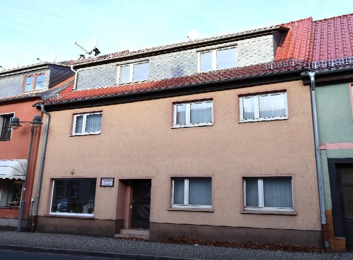 Vorderansicht eines zweistöckigen Wohn- und Geschäftshauses mit brauner Fassade, Eingangstür und Fenstern, Dach mit roten Ziegeln und Satellitenschüssel