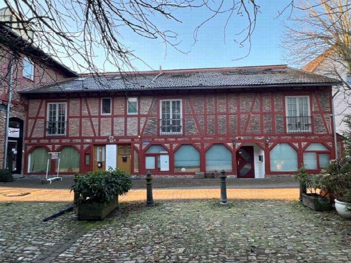 Vorderansicht eines zweistöckigen Fachwerkhauses mit roten Holzbalken und Ziegeldach, Eingangstüren und Fenstern, gepflastertem Vorplatz und Pflanzenkästen