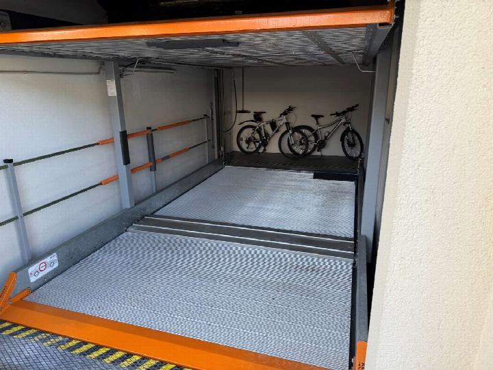Mehrfachsteg-Parkplatz mit Metallboden und orangefarbenen Sicherheitsleitern, im Hintergrund zwei Fahrräder auf oberer Ebene