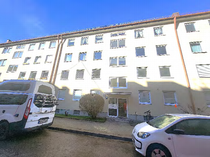 Eingangsbereich eines mehrstöckigen Wohnhauses mit weißer Fassade, Eingangstür, Fenstern und geparkten Fahrzeugen vor dem Gebäude