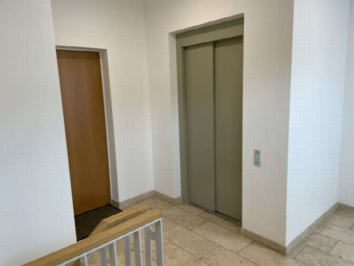Treppenhaus mit Aufzugstür und Holztür, Fliesenboden, Geländer im Vordergrund