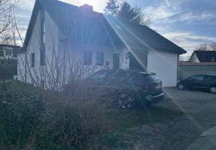 Seitenansicht eines Einfamilienhauses mit angrenzendem Kiesplatz und geparkten Fahrzeugen, teilweise verdeckt durch Sträucher