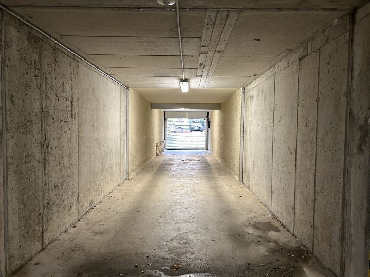 Unterirdischer Parkplatzgang mit Betonwänden und -boden, beleuchtet durch Deckenleuchten, führt zu einer Rolltoröffnung mit Blick auf geparkte Fahrzeuge