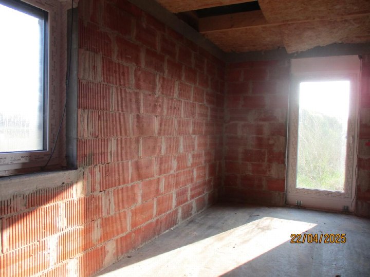 Unfertiger Innenraum mit roten Ziegelwänden, Betonboden und Holzdeckenbalken, zwei Fenstern, Tageslicht