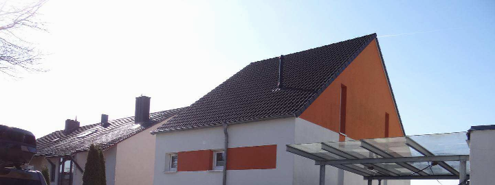 Seitenansicht eines Einfamilienhauses mit schwarzem Ziegeldach, weißer und orangefarbener Fassade sowie einem Glasdachüberstand