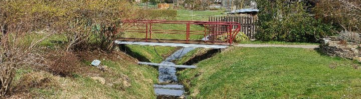 Gartenbereich mit rotem Metallbrückchen über einem kleinen Bach, grünen Wiesen und Sträuchern im Vordergrund