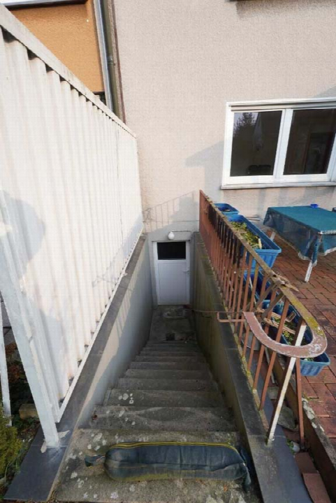 Außenansicht einer Betontreppe mit Geländer, die zu einer weißen Tür im Erdgeschoss führt, neben einem gepflasterten Terrassenbereich