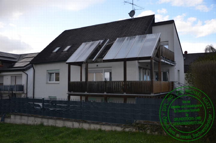 Zweistöckiges Reihenhaus mit dunklem Satteldach, Balkonen und Holzveranda; vorderer Teil des Grundstücks mit Metallzaun und Grünfläche im Vordergrund