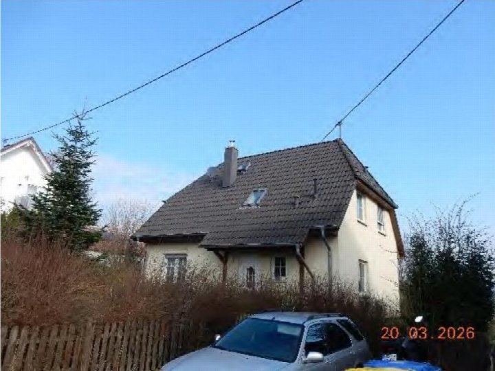 Einfamilienhaus mit Satteldach und hellen Wänden, vorne gesehen, umgeben von Hecken und einem Holzzaun, ein Auto im Vordergrund
