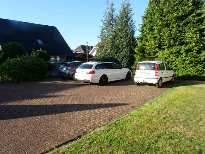 Pflastersteine gepflasterte Einfahrt mit drei geparkten Autos vor einem Wohnhaus mit dunklem Dach und grüner Umgebung