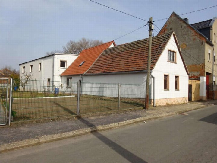 Seitenansicht eines weißen Gebäudes mit rotem Ziegeldach in Teuchern OT Nessa, Kapellenende 69, hinter einem Metallzaun und neben einer Straße