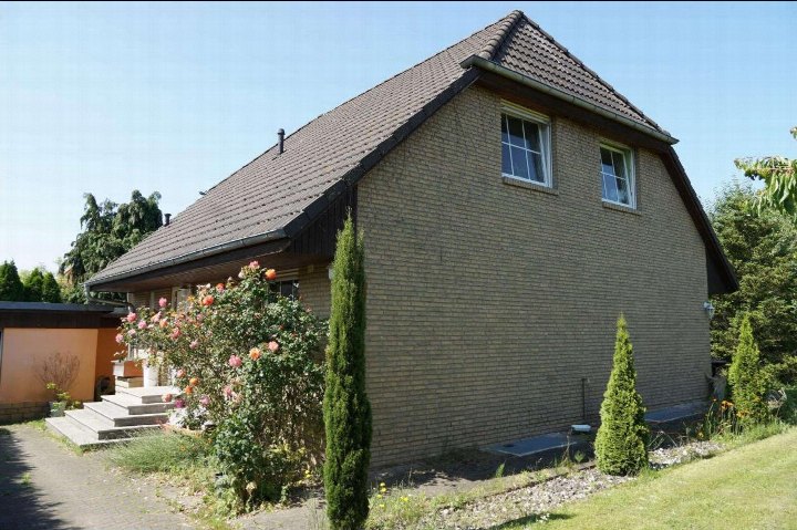 Seitenansicht eines Einfamilienhauses mit Ziegelfassade, Satteldach und Gartenbeeten mit Rosen und Nadelbäumen