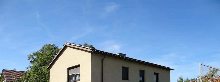 Seitenansicht eines beigefarbenen Gebäudes mit Dach, Fenstern und umgebenden Bäumen unter blauem Himmel
