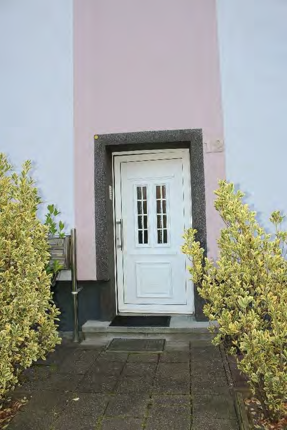 Eingangstür eines Gebäudes mit weißer Tür, dunklem Rahmen und beidseitigen Sträuchern. Garagenzugang