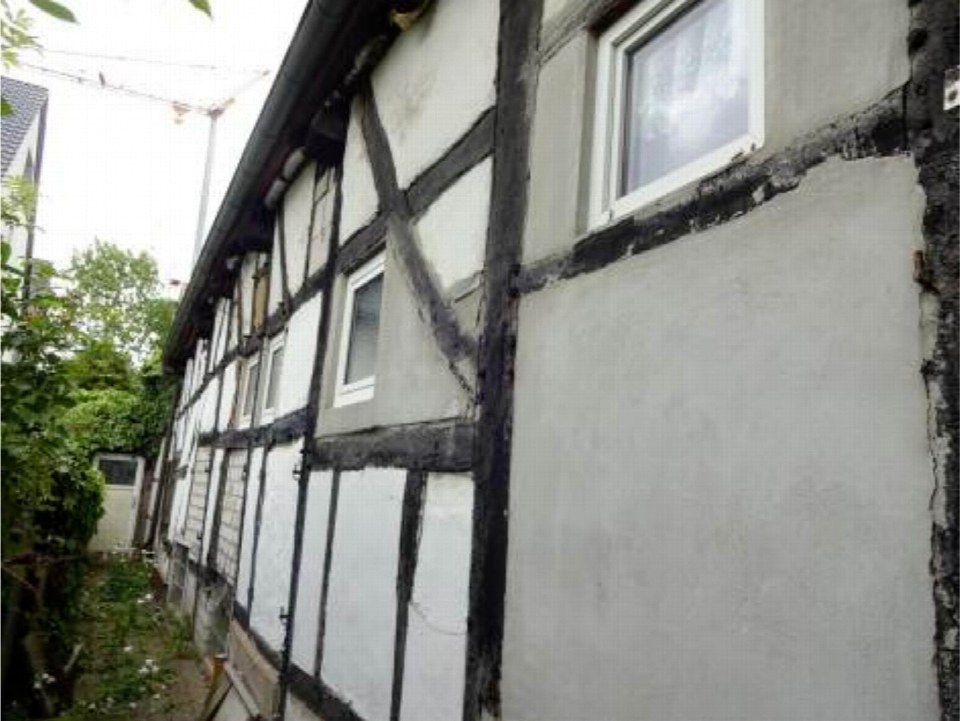 Seitenansicht eines Fachwerkhauses mit weißen Wänden und dunklen Holzbalken, Fenstern und angrenzender Grünfläche