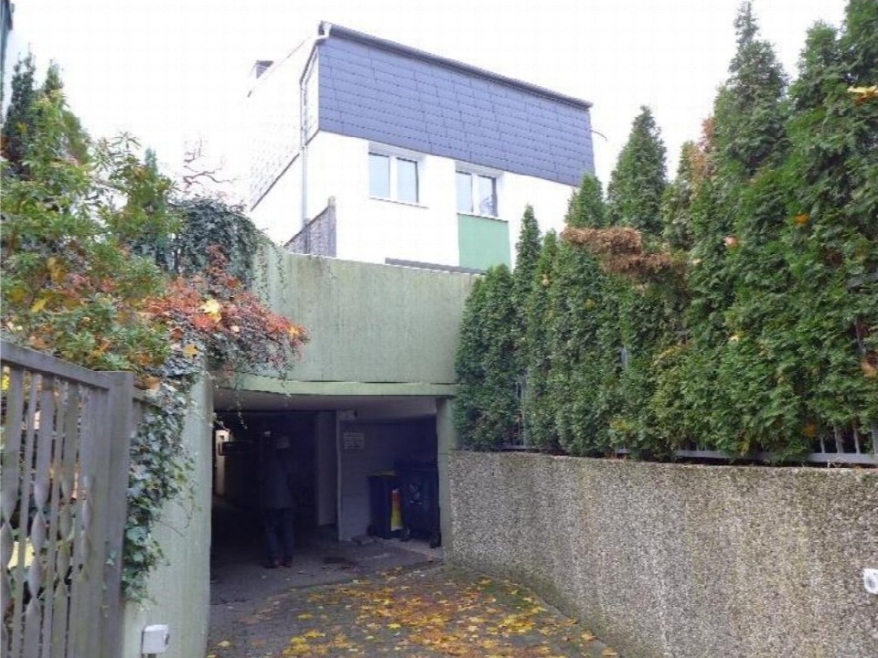 Eingangsbereich eines Reihenhauses mit Betonüberdachung, Gittertor und umgebenden Hecken. Ein Mensch betritt den unterirdischen Bereich