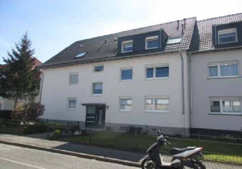 Fassade eines Mehrfamilienhauses mit Dachgauben, Fenstern und Eingangsbereich, vorne gesehen, mit gepflastertem Gehweg und einem Elektroroller im Vordergrund