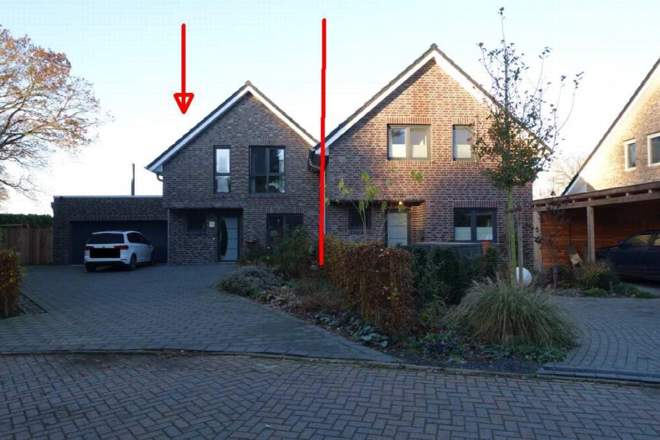 Vorderansicht einer Doppelhaushälfte mit Ziegelwand, Dachgauben, Einfahrt und Garage. Im Vorgarten sind Sträucher und ein Baum zu sehen