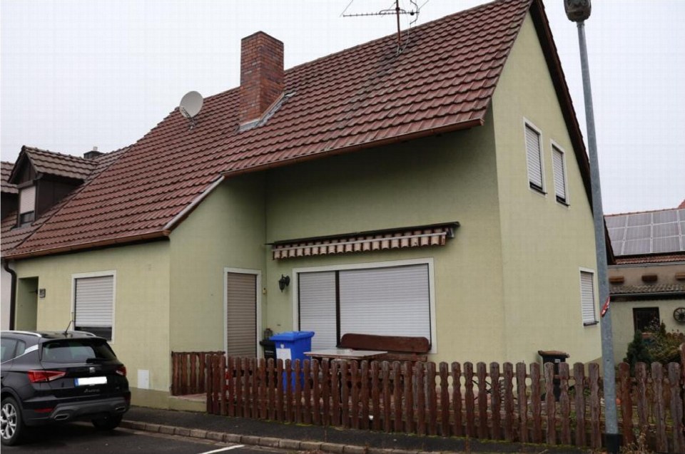 Seitenansicht eines Einfamilienhauses mit braunem Ziegeldach, hellgrüner Fassade, Fenstern mit Jalousien und einem Holzzaun im Vorgarten