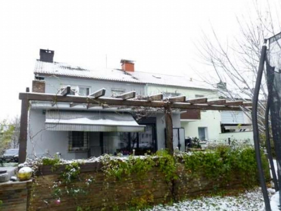 Gartenbereich eines Reihenhauses mit Holzpergola, schneebedecktem Boden und grünen Sträuchern vor einer Mauer