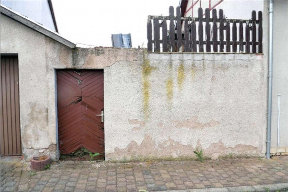 Eingangstür aus rotem Holz in einer verwitterten Betonmauer, gepflasterter Weg davor, kleiner Holzzaun oben