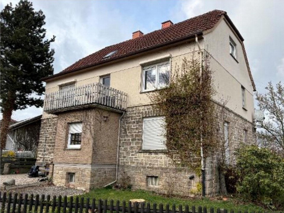 Seitenansicht eines Einfamilienhauses mit Steinsockel, Stuckfassade, Dachgauben und Balkon, umgeben von Grünanlagen und einem Zaun