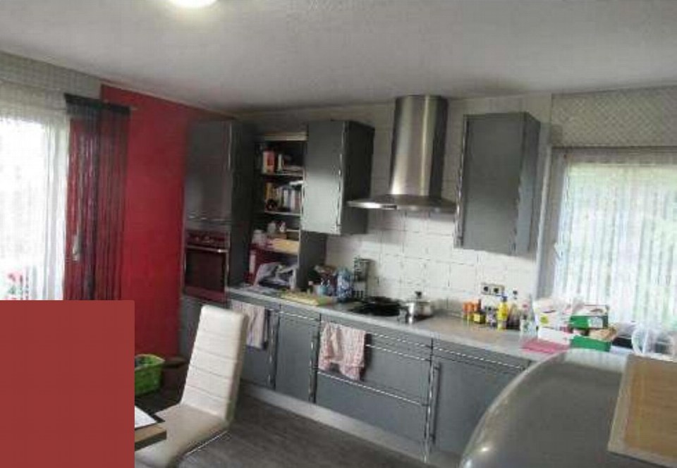 Küche mit grauen Schränken, Edelstahl-Abluftkamin und Arbeitsfläche, Fenster mit Vorhängen, rote Wand im Hintergrund