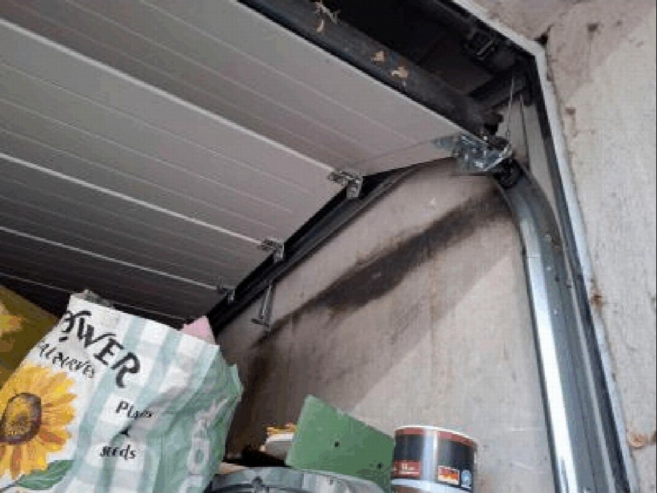 Innenansicht einer Garage mit teilweise geöffneter Rolltür, Metallführungen und aufbewahrten Gegenständen wie einem Sack und einer Dose