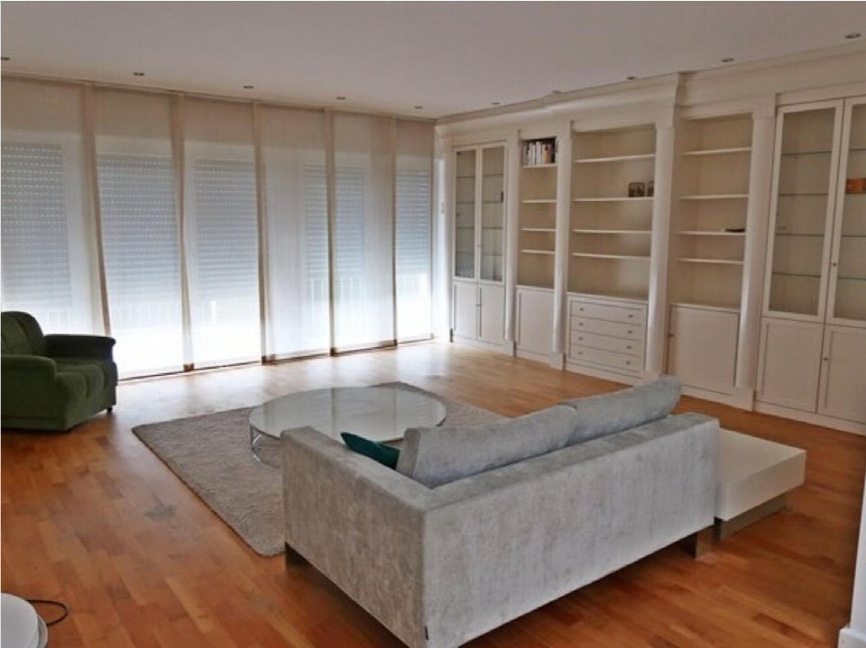 Wohnzimmer mit grauem Sofa, grünem Sessel, runder Glas-Tisch, Holzboden und großen Fenstern mit Jalousien. Einbauschränke an der Wand